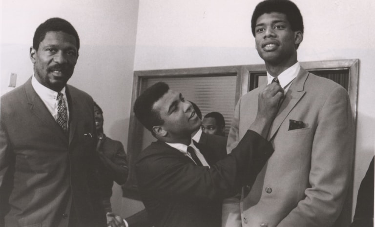 Black and white photo of Muhammad Ali in a suit, fixing the tie of another man in a suit, standing