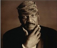 Jim Brown, wearing black jacket, white shirt and kufi hat poses with hand to his chin