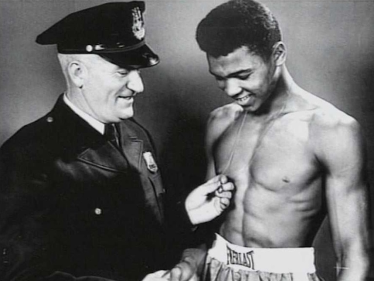 Black and white photo of a young Muhammad Ali shirtless in boxing shorts with Joe Martin in a police uniform