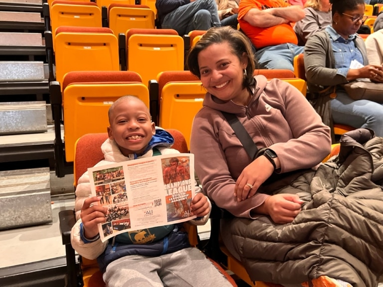 Parent and child sitting in auditorium-style seats as the child smiles and holds up a "Champions League Youth Leadership Academy" brochure