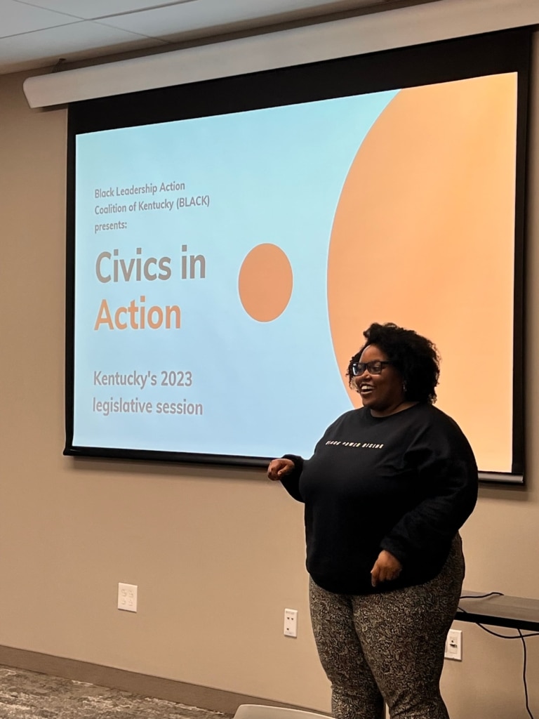A person wearing glasses and black clothing stands next to a projection screen for "Civics in Action"
