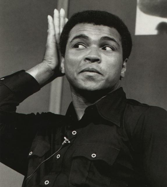 Muhammad Ali pats his head and looks to the side with a proud expression