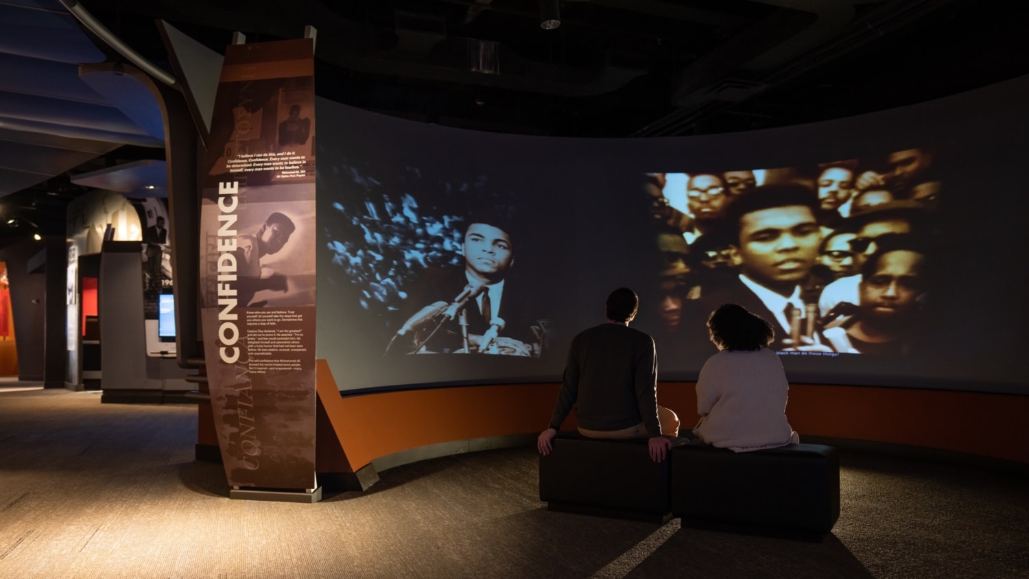 Two people sitting in a dark exhibit gallery watching a video about Muhammad Ali on a large projection screen