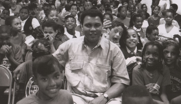 Black and white photo of Muhammad Ali sitting in a room full of students