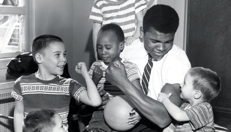Black and white photo of Muhammad Ali in a shirt and tie with a number of young children in leg braces