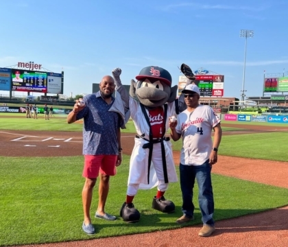 Two men pose with a bat mascot on a baseball field