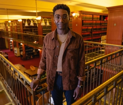 Photo of man wearing glasses and brown jacket, sweater and jeans standing in front of bookcases at library