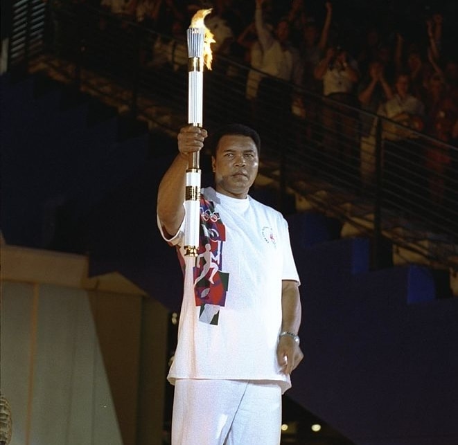 Muhammad Ali dressed in all white lifts up a lit Olympic Torch