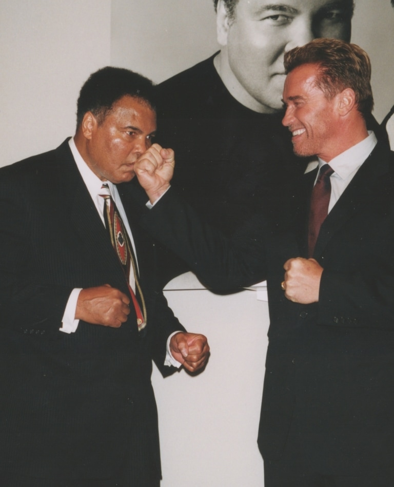 Muhammad Ali and Arnold Schwarzenegger posing for camera as Arnold holds punch toward Ali