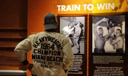 Person with Muhammad Ali jacket and baseball cap reading an exhibit wall display