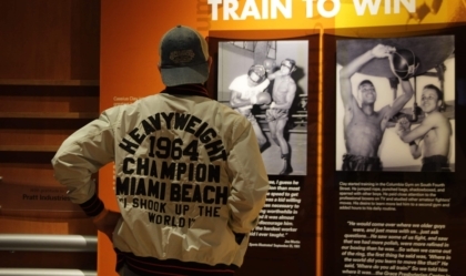 Person with Muhammad Ali jacket and baseball cap reading an exhibit wall display