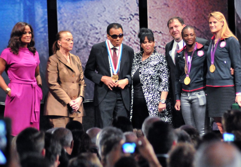 Photo of Muhammad Ali wearing medal surrounded by group on stage while audience takes pictures