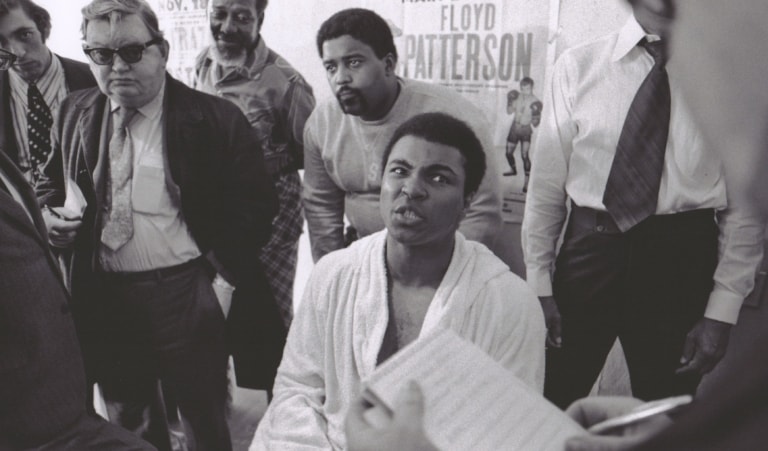 Muhammad Ali snarls at a reporter while surrounded by men.