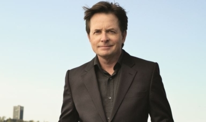 Michael J. Fox posing with black suit and black shirt in front of body of water