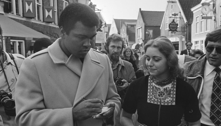 Black and white photo of Muhammad Ali in a coat, signing an autograph book for a woman in traditional Dutch dress surrounded by several photographers