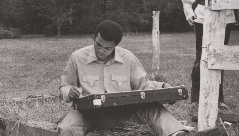 Black and white photo of Muhammad Ali as a young man, seated, drawing outdoors while another man stands nearby and looks at his work