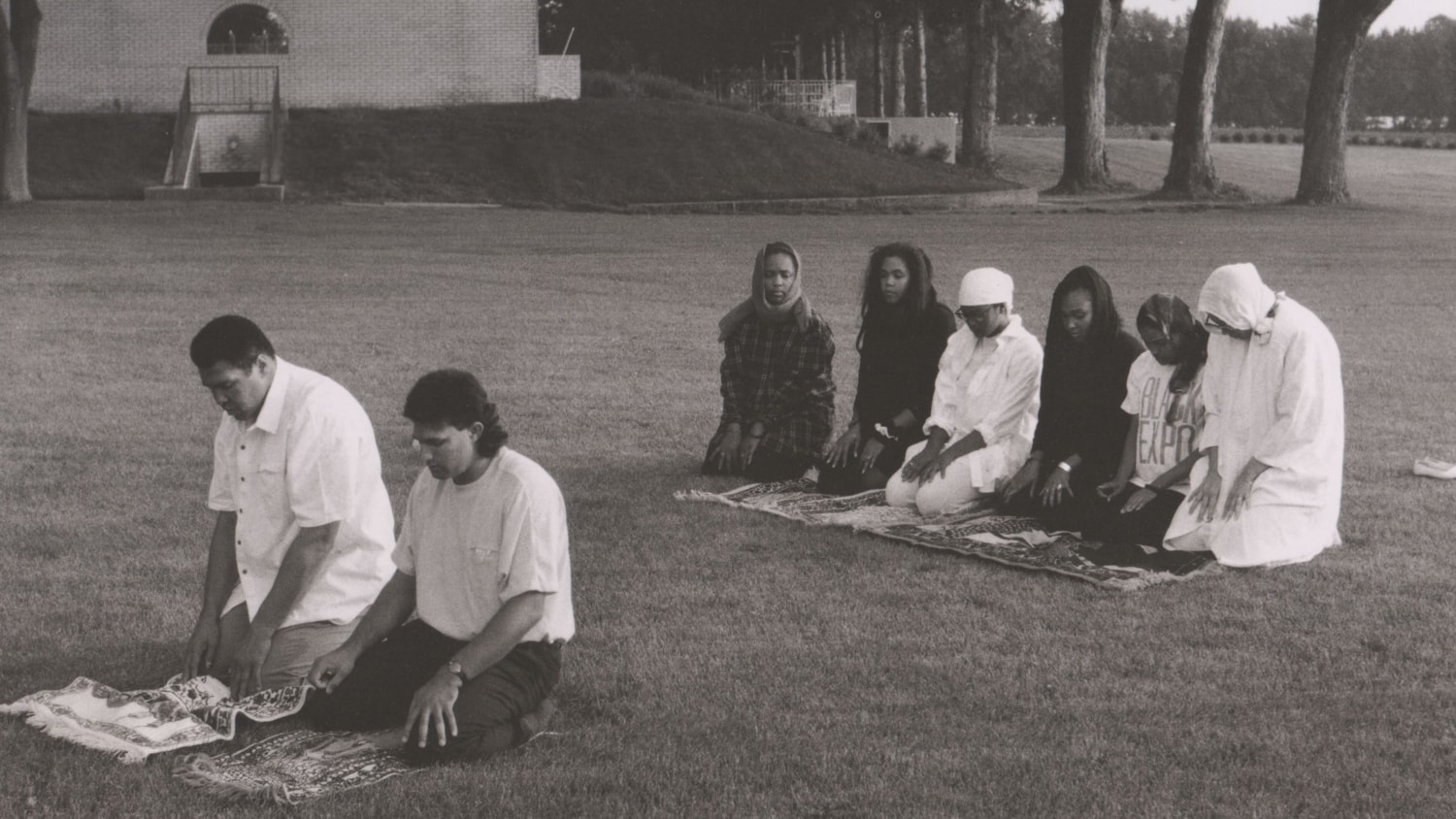 Black and white photo of Muhammad Ali with family and guests kneeling and praying on a lawn