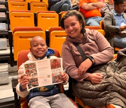 Parent and child sitting in auditorium-style seats as the child smiles and holds up a "Champions League Youth Leadership Academy" brochure