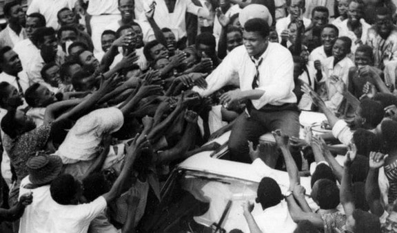 Muhammad Ali in Nigeria sitting on top of a car greeting a huge crowd of people in 1964.