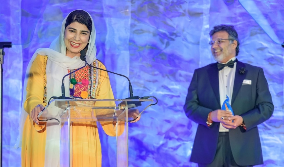 Woman in yellow outfit wearing hijab making speech at lecturn while man in suit holds award to her right