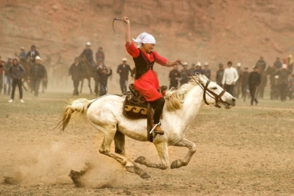Photo of woman wearing red blouse, headscarf and black pants riding a white horse