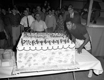 Black and white photo of Muhammad Ali blowing out candles on big birthday cake