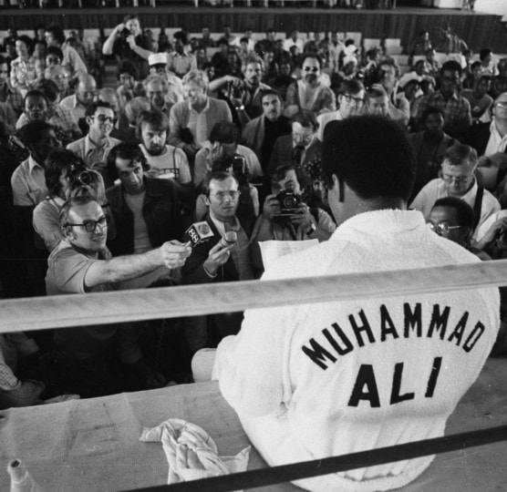 Vintage photo of Muhammad Ali sitting on the outside edge of a boxing ring speaking to a group of reporters