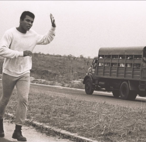 Black and white photo of Muhammad Ali running in sweats while waiving to a large truck full of people waving at him