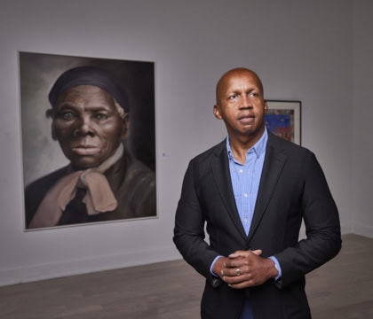 Man wearing dark jacket and blue button-up shirt inside room filled with museum art looking off camera