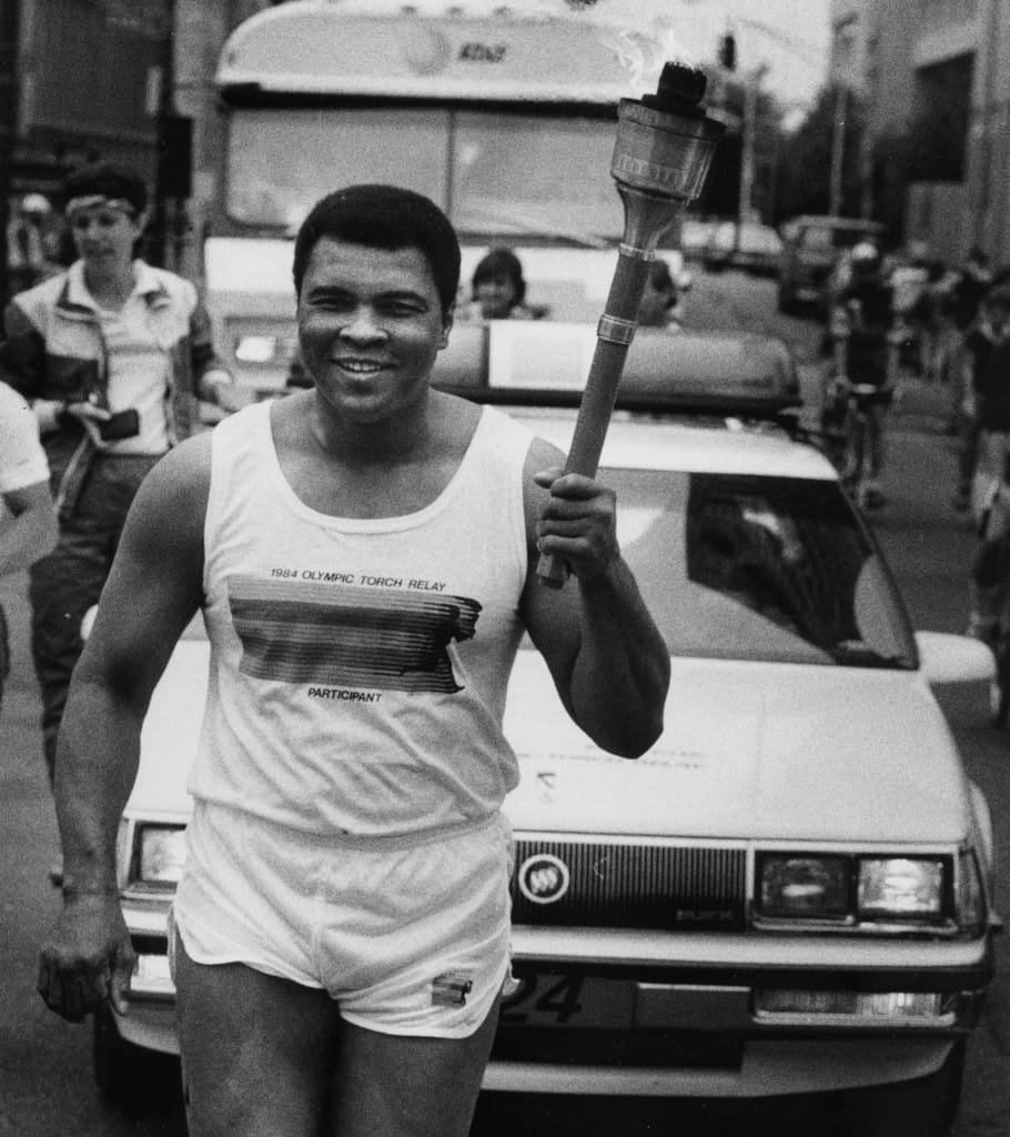 Black and white photo of Muhammad Ali running with the Olympic Torch in the middle of a street