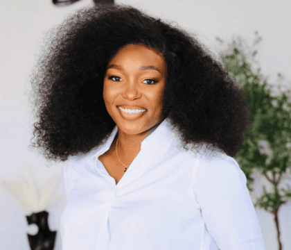 Photo of woman with long hair wearing a white button-up shirt and dark pants smiling at camera