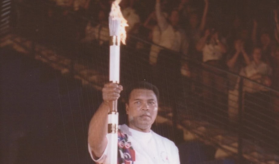 Photo of Muhammad Ali wearing white shirt and pants holding Olympic torch