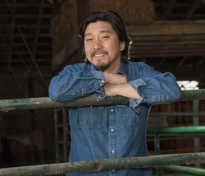 Picture of man with long hair and beard wearing denim shirt leaning over railing and smiling at camera