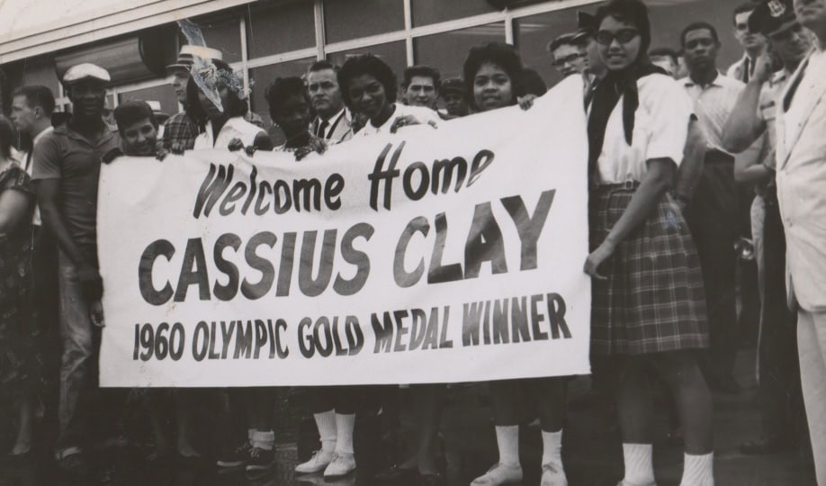 Group of people holding banner that says "Welcome Home Cassius Clay"