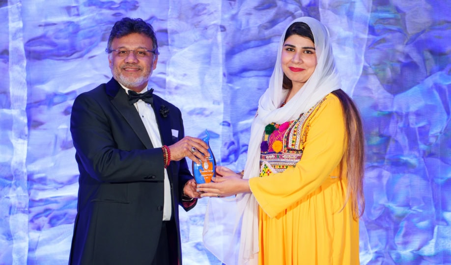 Woman in yellow outfit wearing hijab accepts award from man in suit