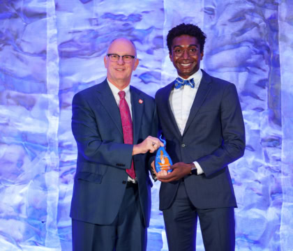 Photo of man in blue suit and pink tie and young man wearing dark suit and blue bow tie posing and holding award