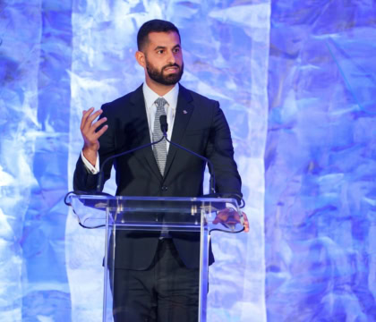 Man with short hair and beard wearing dark suit speaks at podium