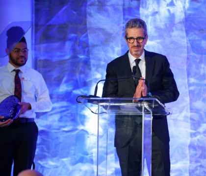 Man wearing dark suit and black glasses speaking at podium with docent in white shirt and tie standing behind