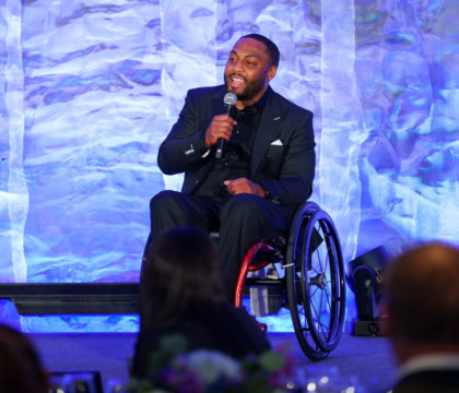 Man in dark suit and tie sitting in wheelchair speaking in a microphone