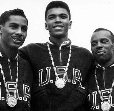 Black and white photo of Muhammad Ali wearing USA jacket and gold medal alongside two men facing camera