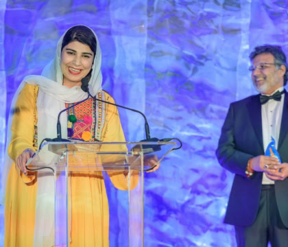 Woman in yellow outfit wearing hijab making speech at lecturn while man in suit holds award to her right