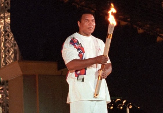 Photo of Muhammad Ali wearing white shirt and pants holding Olympic torch