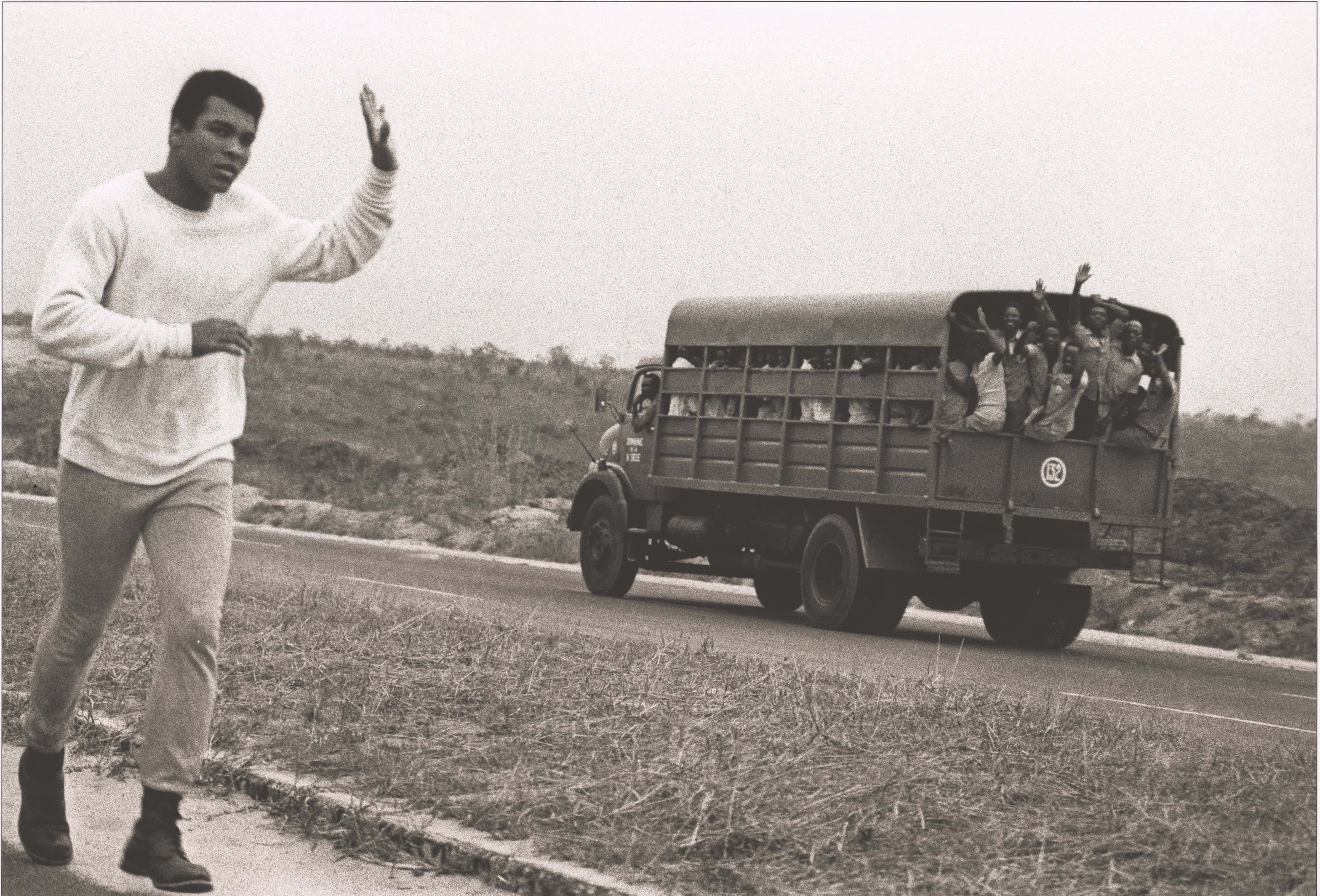 Black and white photo of Muhammad Ali running and waving to passing bus