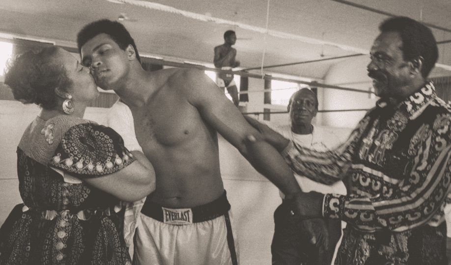 Black and white photo of Muhammad Ali with his mother and father as he leans in for a kiss from his mother