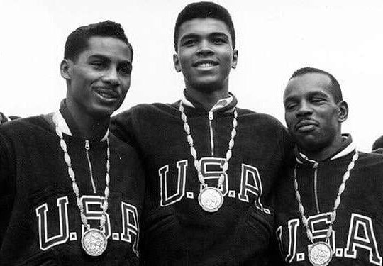 Black and white photo of Muhammad Ali wearing USA jacket and gold medal alongside two men facing camera