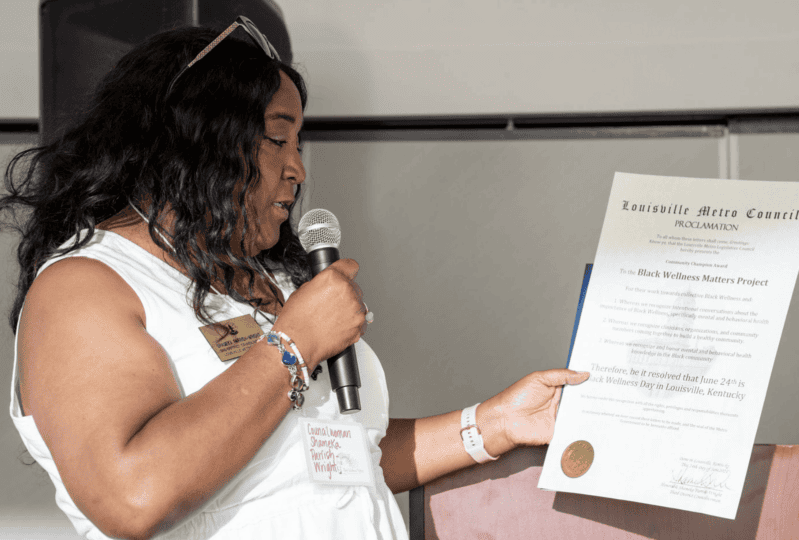 Woman wearing white top holding proclamation while speaking at microphone