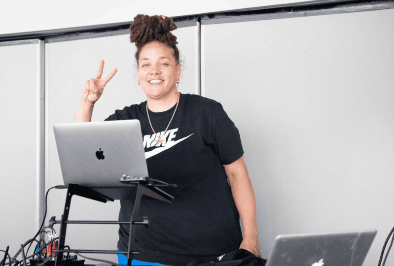 Woman at table with multiple laptops smiling and making peace sign at camera