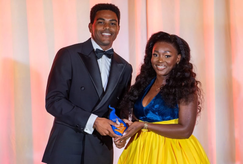 Photo of man in black suit and woman in blue and yellow dress holding award