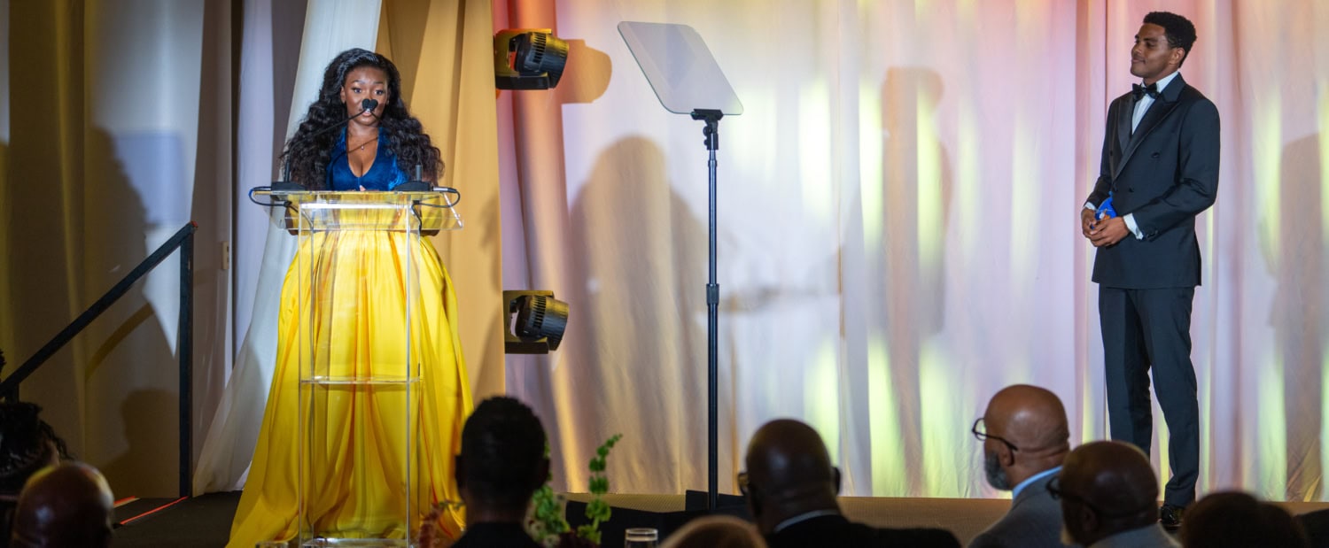 Photo of woman standing at podium wearing blue and yellow dress as man in suit looks toward her