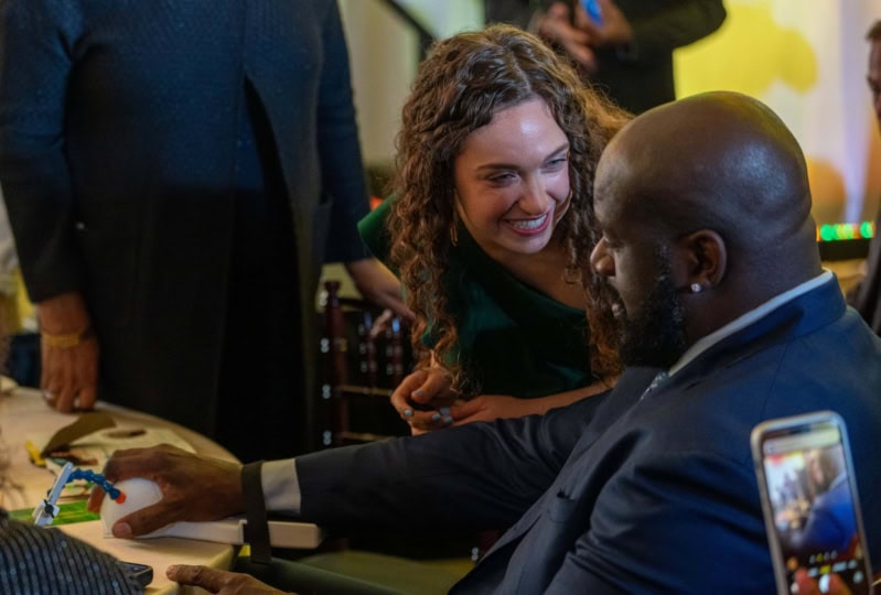 Photo of woman with dark curly hair showing Shaquille O'Neal a prototype "Guided Hands" tool at a table